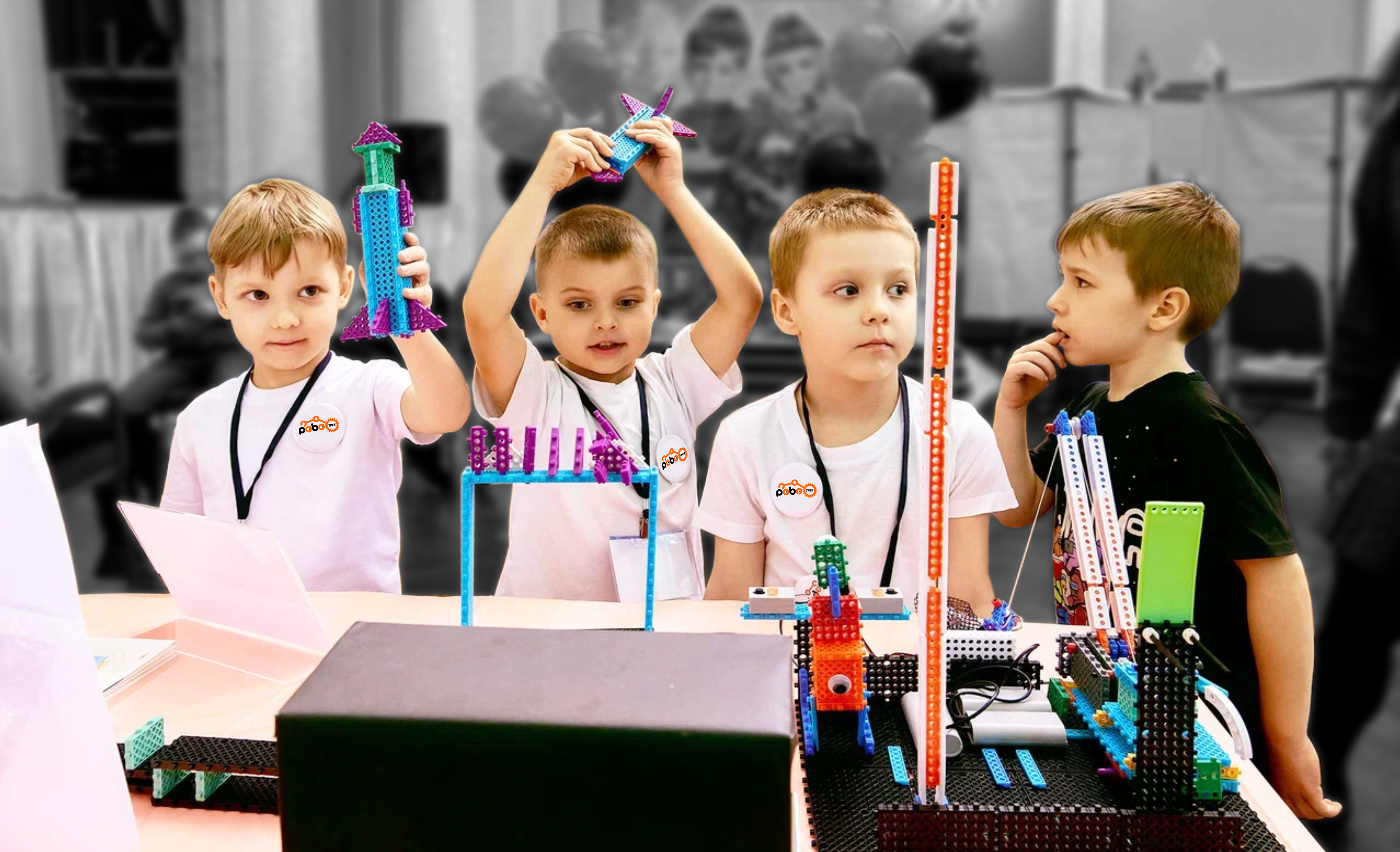 Small robot built from a construction kit standing on a table in a brightly lit room, surrounded by other robotics components and tools, conveying a sense of curiosity and creativity